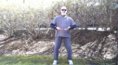 Screenshot image of man, outside, playing T'ai Chi Qigong. His feet are wide and the palms of his hands are drawing upwards, fingertips to fingertips, at his center line.