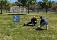 A male facilitator leading a workshop participant in a low, deep hamstring, glute, abductor stretch. 